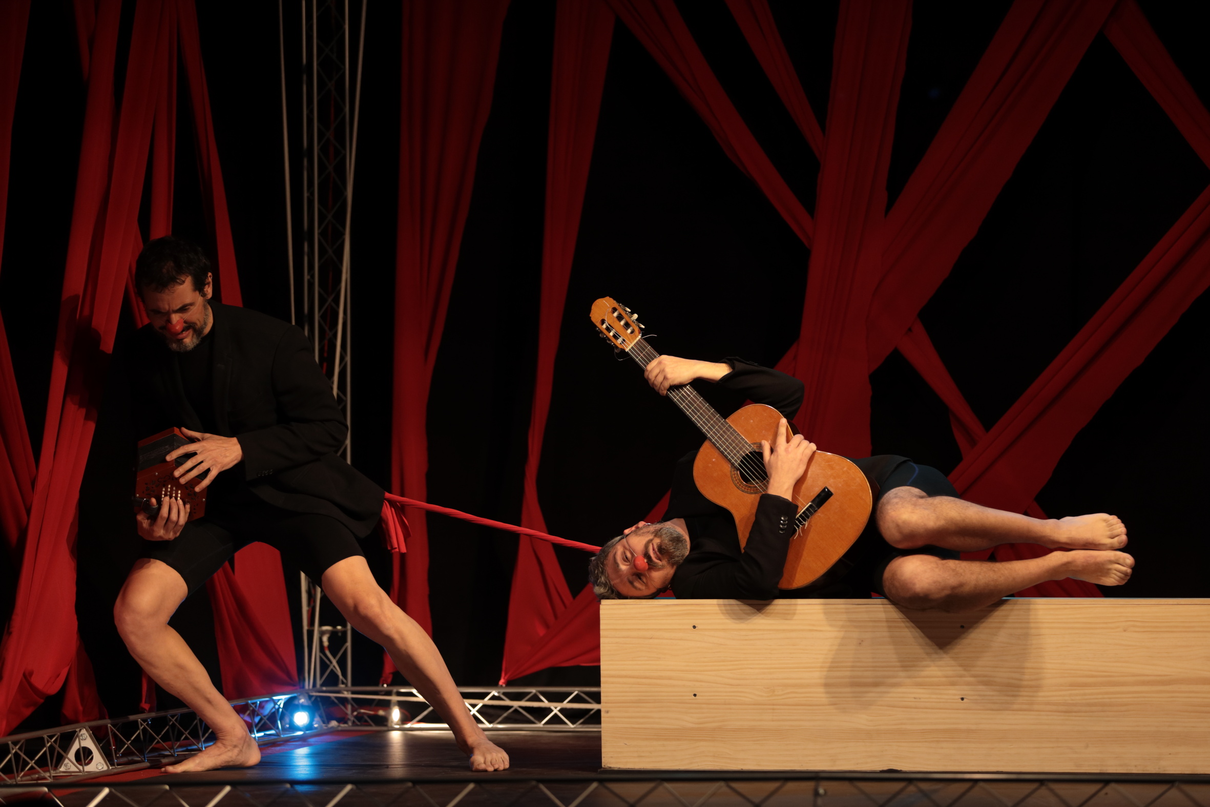 Foto del espectáculo ECO que estuvo en Tarambana. Un hombre con nariz roja está atado a otro que está tumbado en un cajón de madera con una guitarra española. El atado intenta huir.